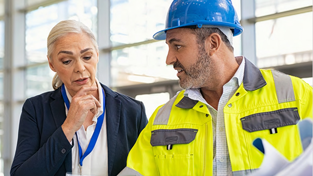 Worker in safety vest and hard hat discusses blueprints with worker in business attire.