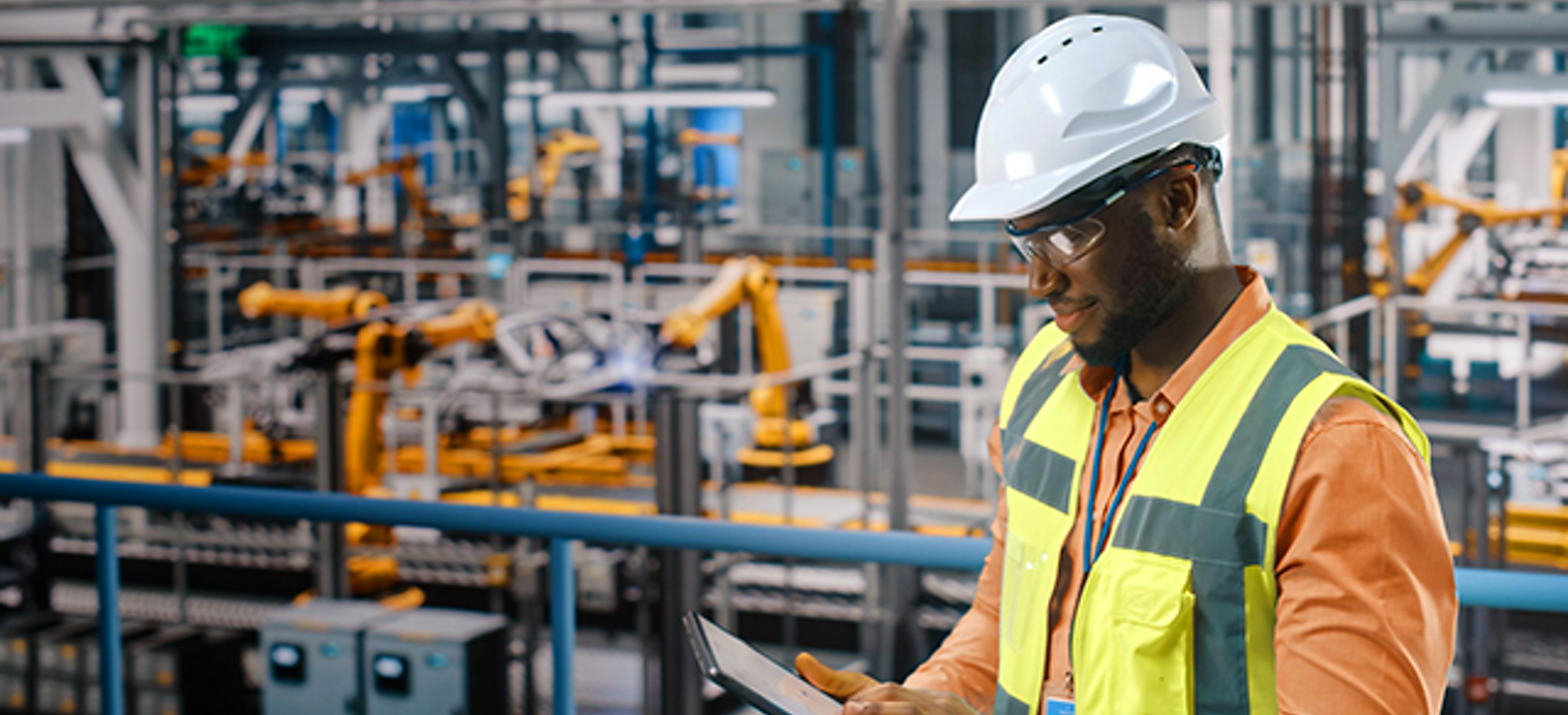 Man in factory setting with hardhat and vest looks down at a tablet.