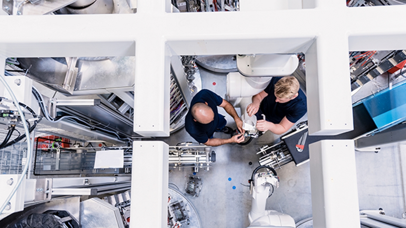 Overhead view of two workers in a manufacturing room with equipment and cables.