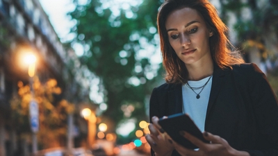 A focused professional checks her smartphone while walking through a city at twilight.