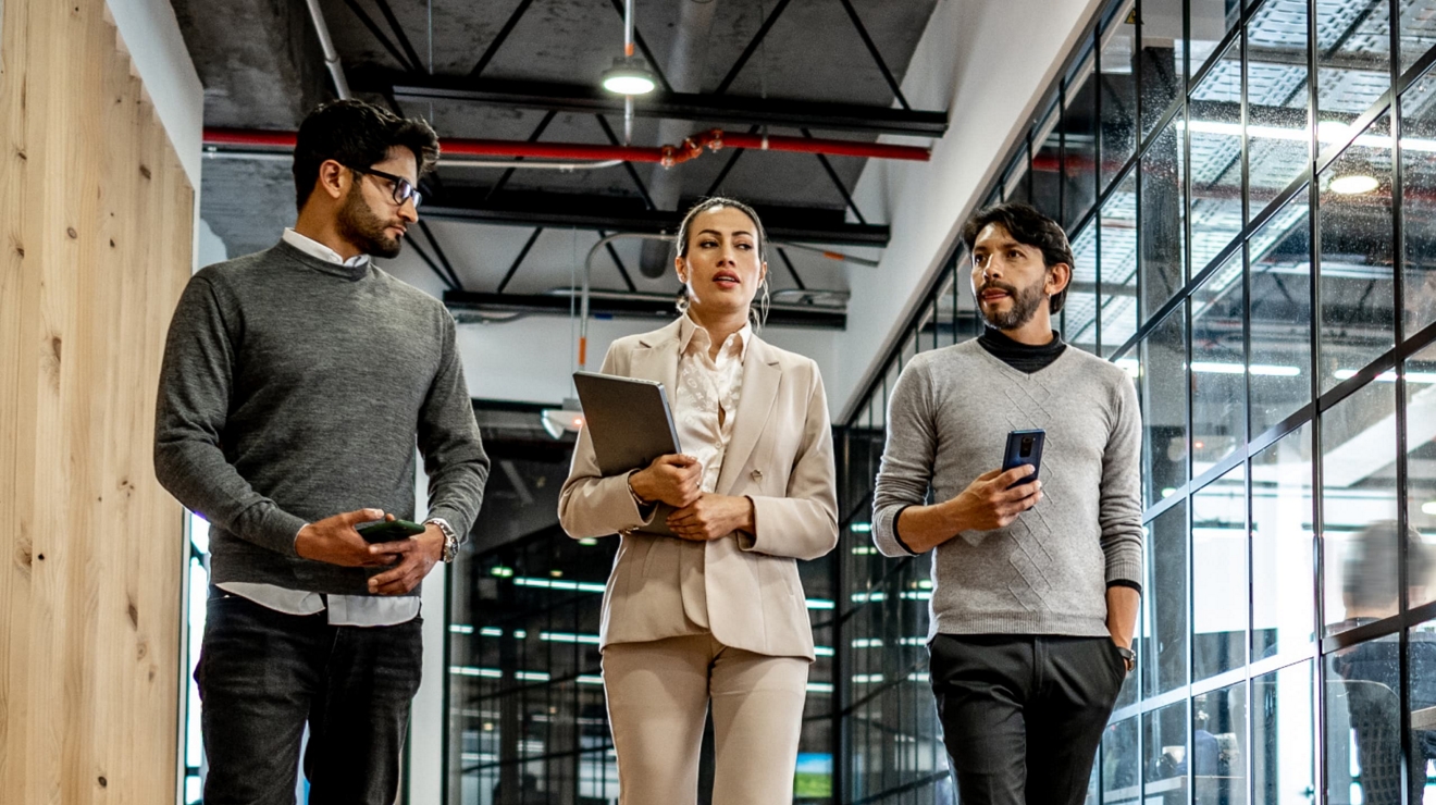 Three colleagues walk through the office and collaborate on the go, each with a mobile device. 