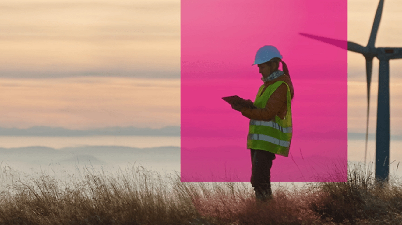 A person works on a network-connected tablet at an open field wearing a hard hat and security vest.