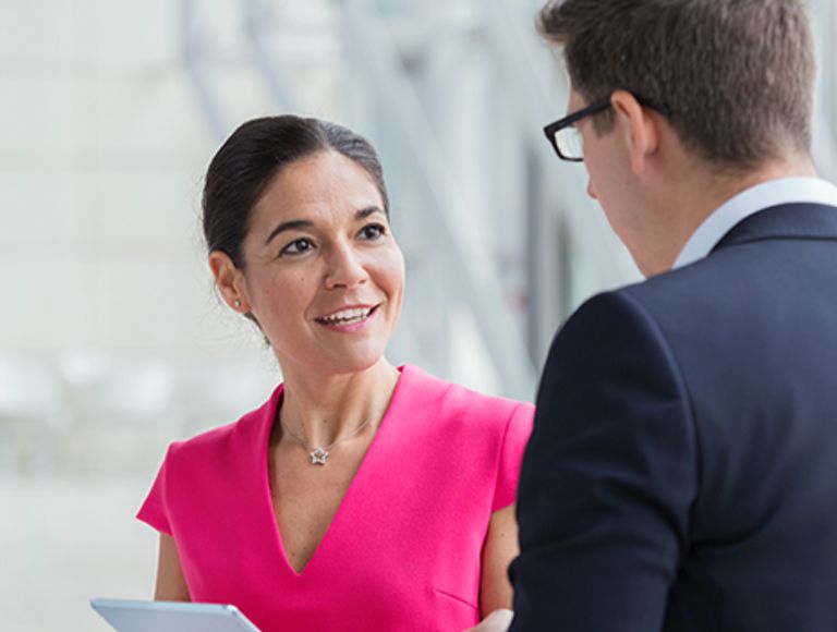 Smiling woman in pink shirt holding tablet while talking to a man in a suit