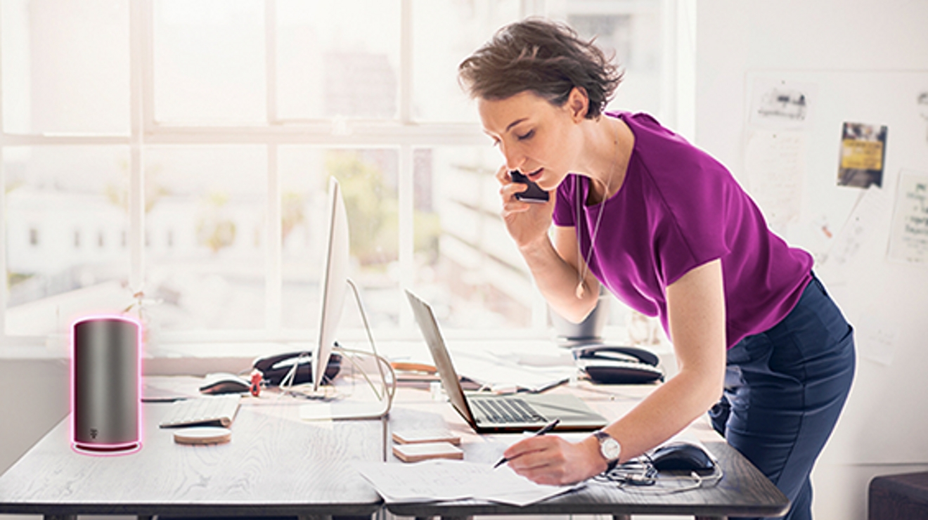 Woman in magenta shirt with smartphone to ear writes on desk with laptop in front of her