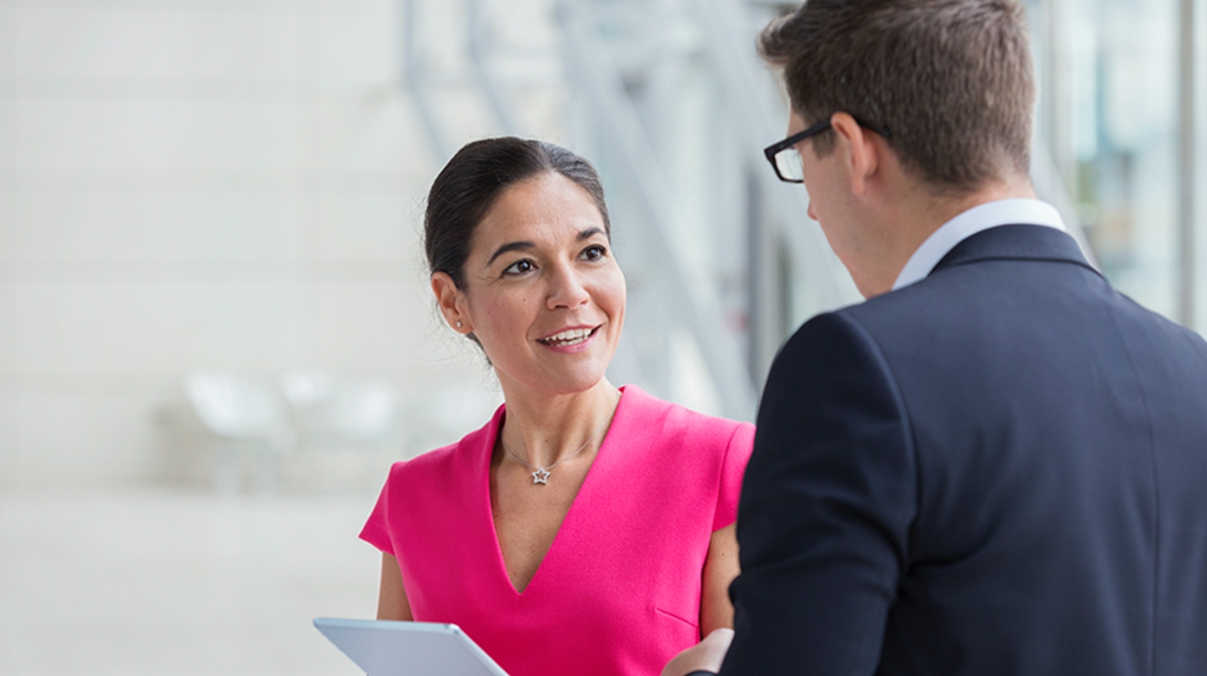 Smiling woman in pink shirt holding tablet while talking to a man in a suit