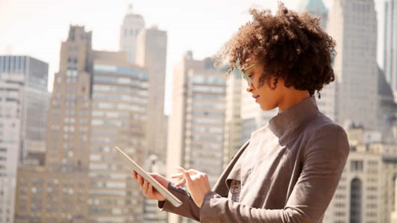 A professional works on a tablet with modern office towers in her background.