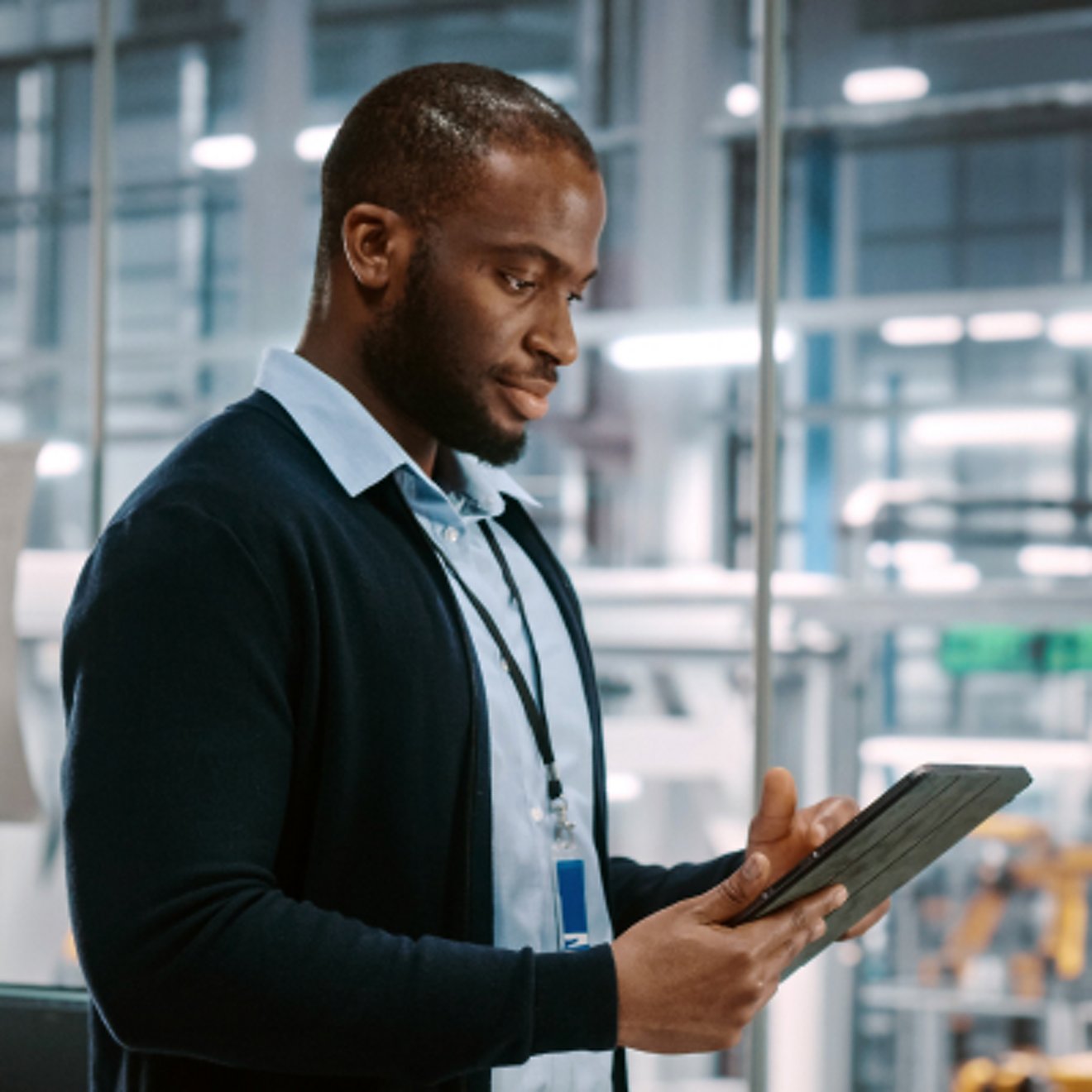 A professional checks a tablet while standing inside an office with view to a manufacturing plant.