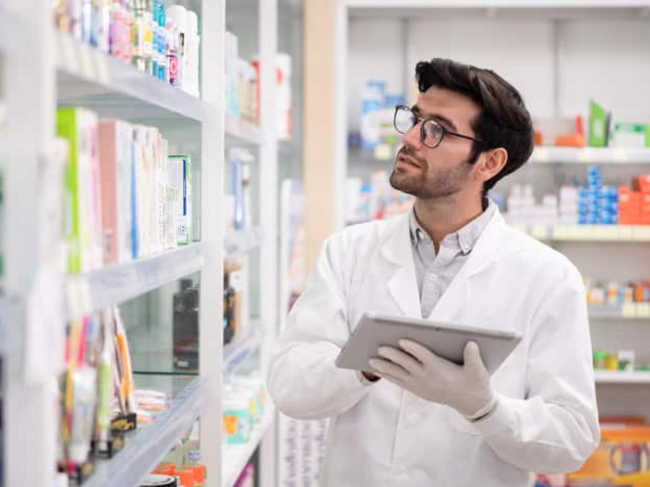 A pharmacist wearing gloves and holding a tablet looks for medication on a shelf.
