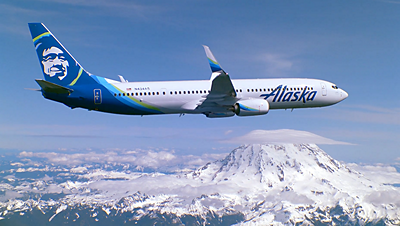 An Alaska Airlines jet flies over the Cascade Mountains in a clear sky.