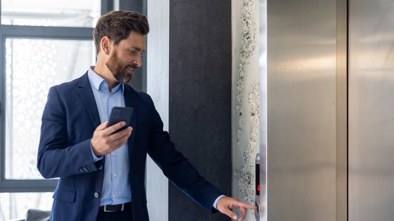 A business executive in a building lobby holds their smartphone as they press an elevator button