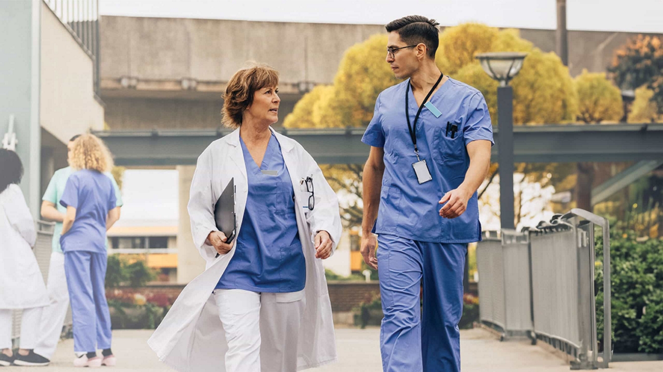 A doctor with a digital tablet under one arm quickly walks along an outdoor path with a colleague.