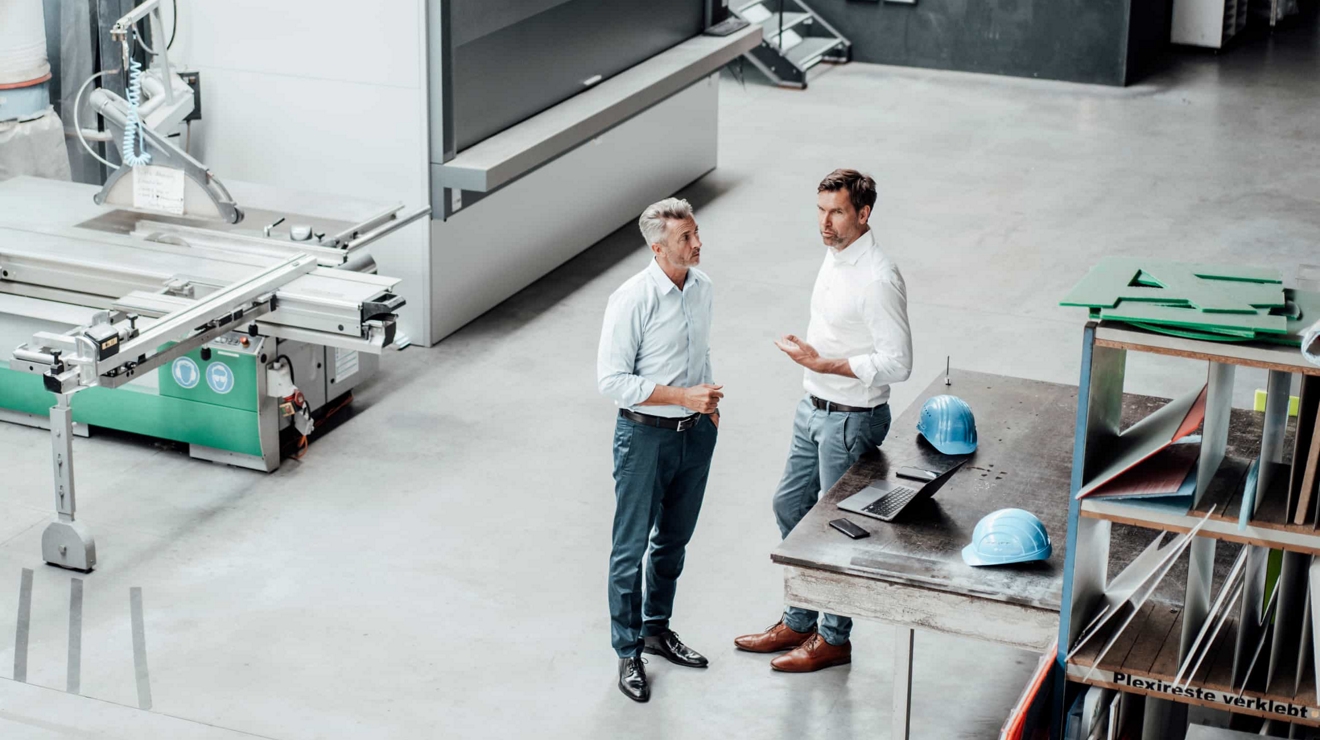 Two colleagues in a manufacturing facility talk beside a table where an open laptop sits. 