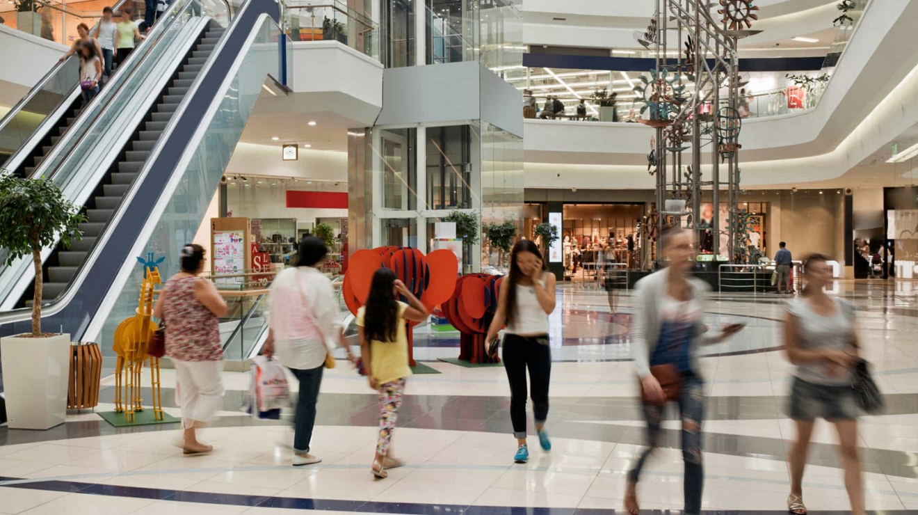 A young adult talks on their cell phone as they walk inside a busy shopping mall.