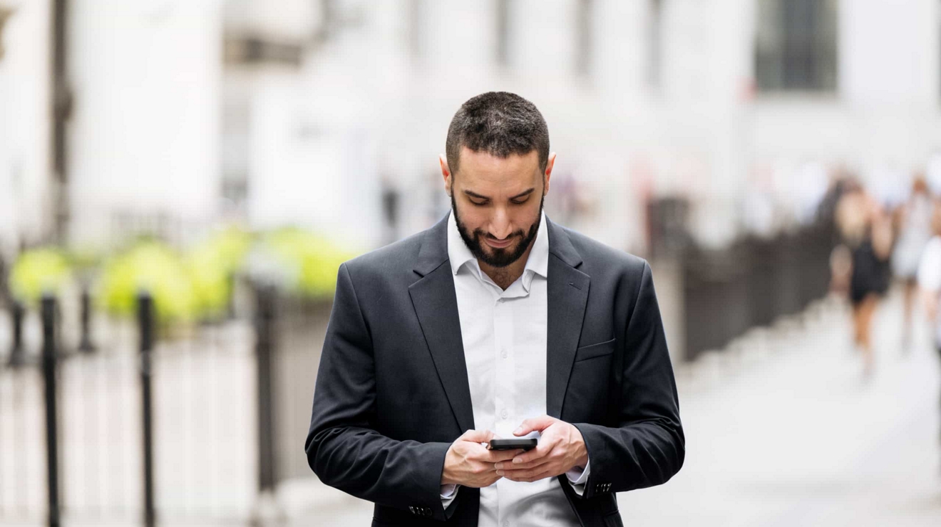 A person in a business suit walks while texting on their connected mobile phone. 