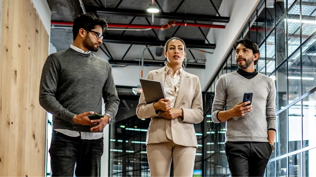 Three coworkers walk and work inside an office holding connected tablets and mobile phones. 