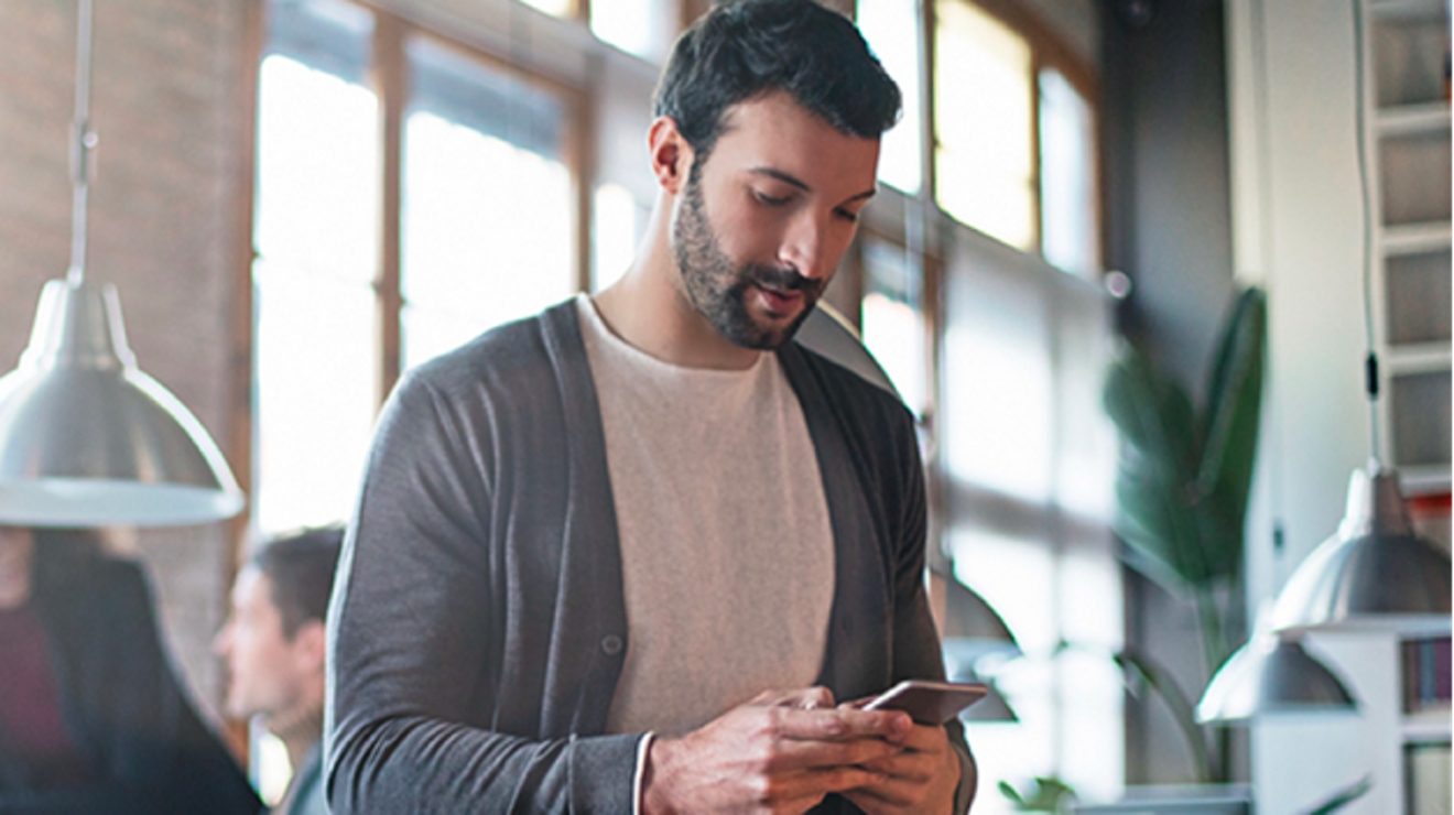 A man in a large airy office area works on his mobile phone while colleagues collaborate behind him.