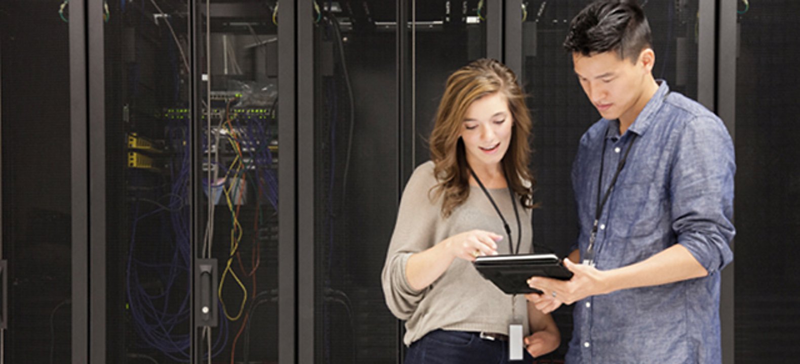 Two people in business casual attire looking at a tablet, while standing in front of office servers.