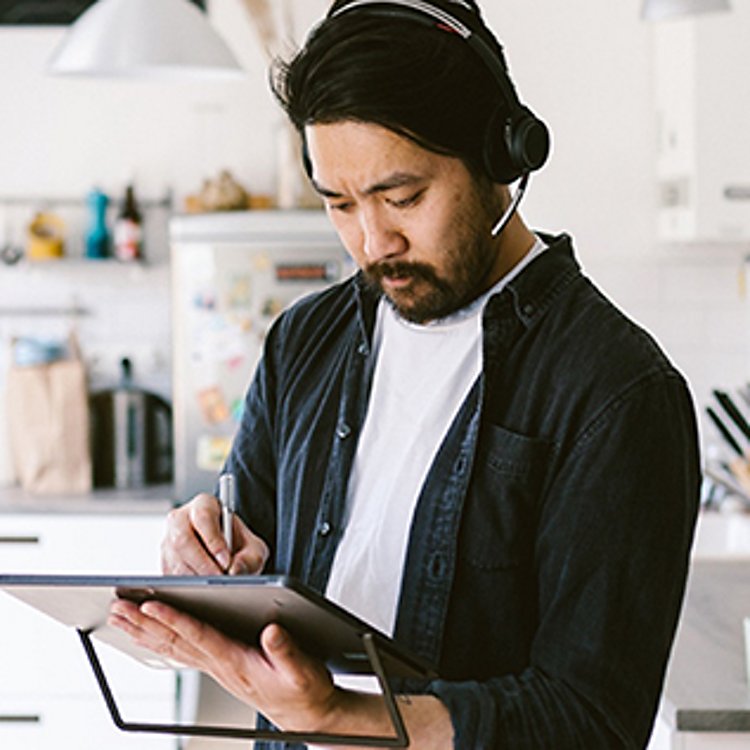 A young man wearing a headset stands in a kitchen and writes on his tablet.