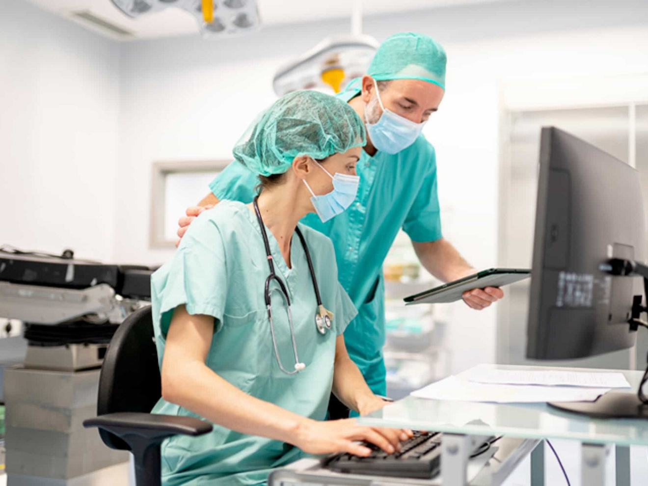 Two doctors in an operating room review data on a desktop computer and a tablet.