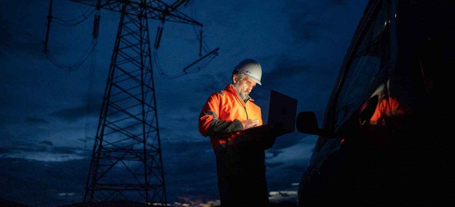 At dusk, a technician stands outside his vehicle and works on a laptop underneath a high-voltage tower.