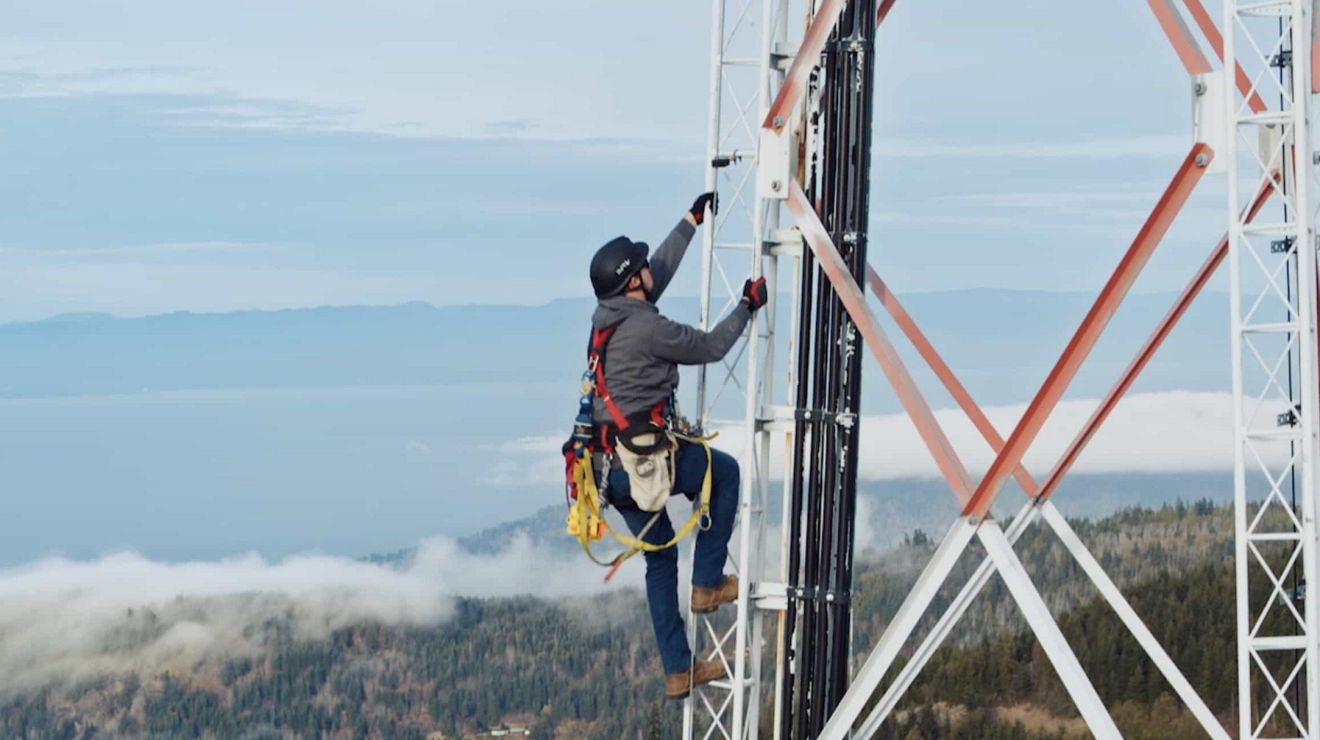 A tower worker wearing safety gear climbs a cell tower in a rural location