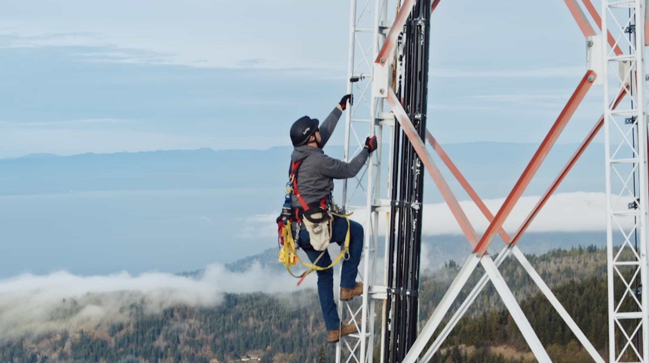 A tower worker wearing safety gear climbs a cell tower in a rural location