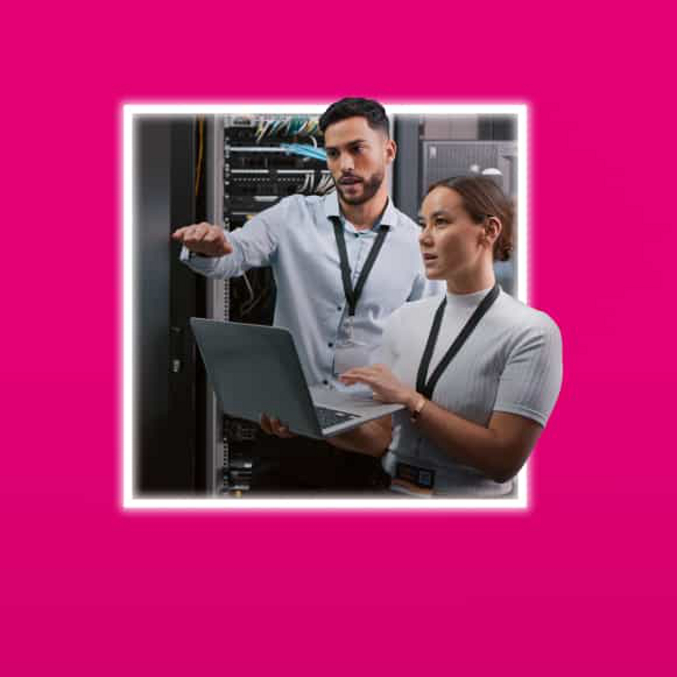 An IT professional holds a laptop and engages in a conversation with a colleague in a server room.