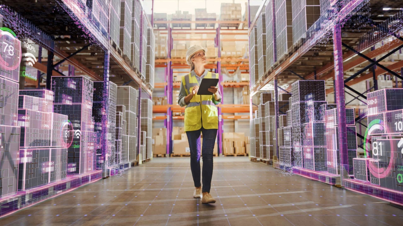 A manager in a hardhat walks a warehouse aisle and checks inventory using a connected tablet. 
