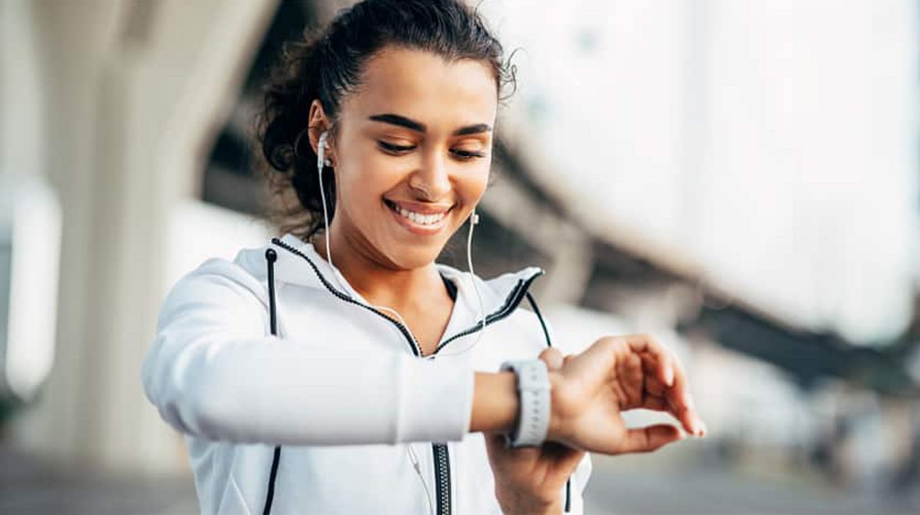 A young woman out for a run listens to earbuds and checks her smartwatch.