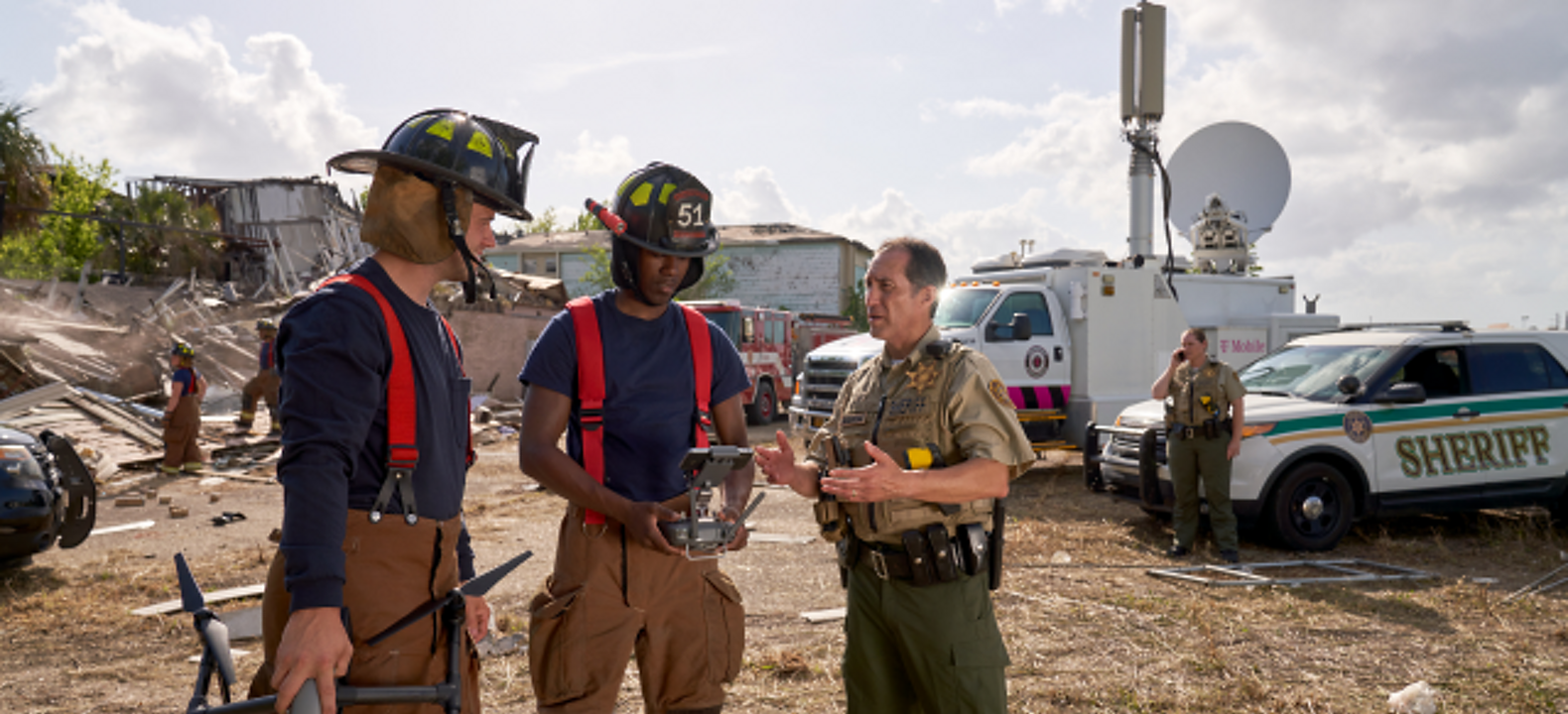 Two firefighters holding a drone and its controller speak with a sheriff at a disaster response site. 
