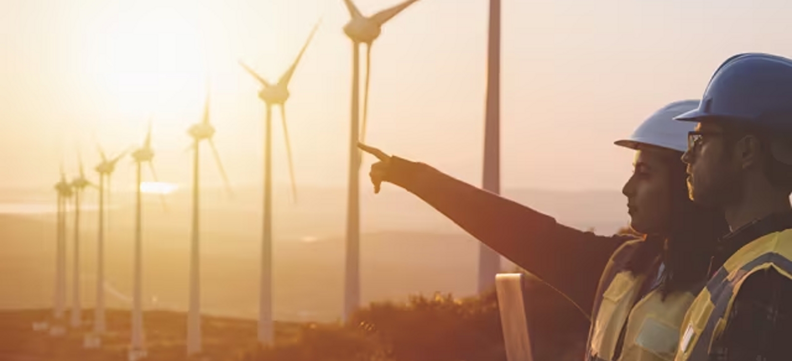 Two engineers, one pointing in the distance and another holding a laptop, at a wind farm at sunrise.