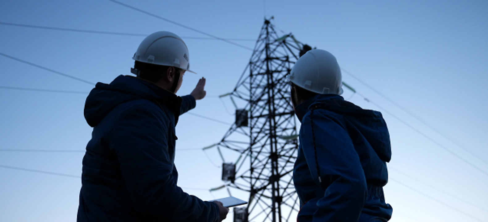 Two energy-utility technicians use a tablet while they evaluate a power-line pylon.