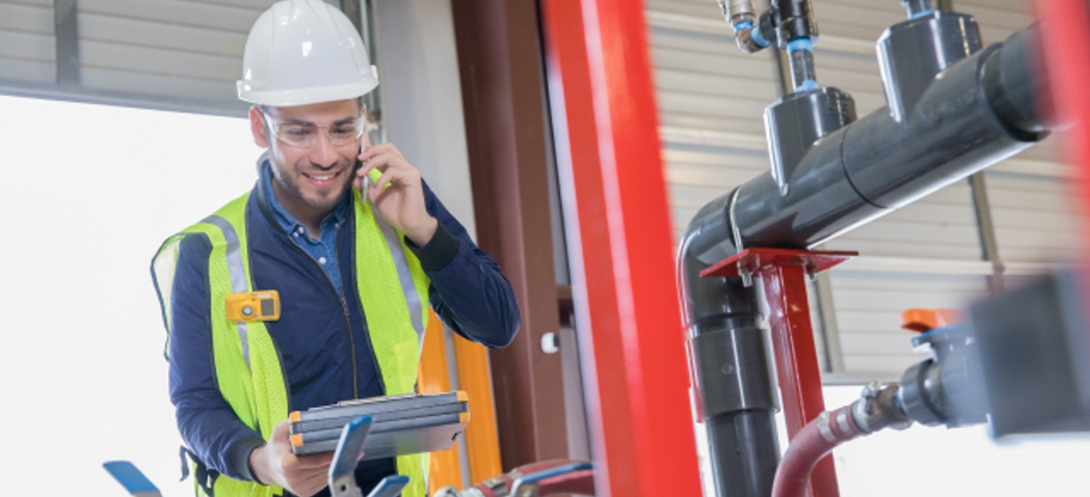Man in overalls with white hard hat stand in front of pipes looking down at a tablet in his hands