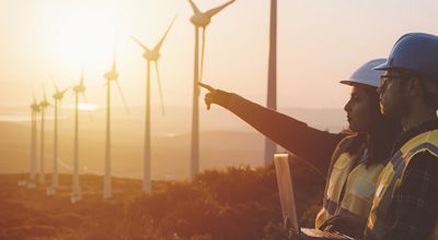 Two engineers, one pointing in the distance and another holding a laptop, at a wind farm at sunrise.
