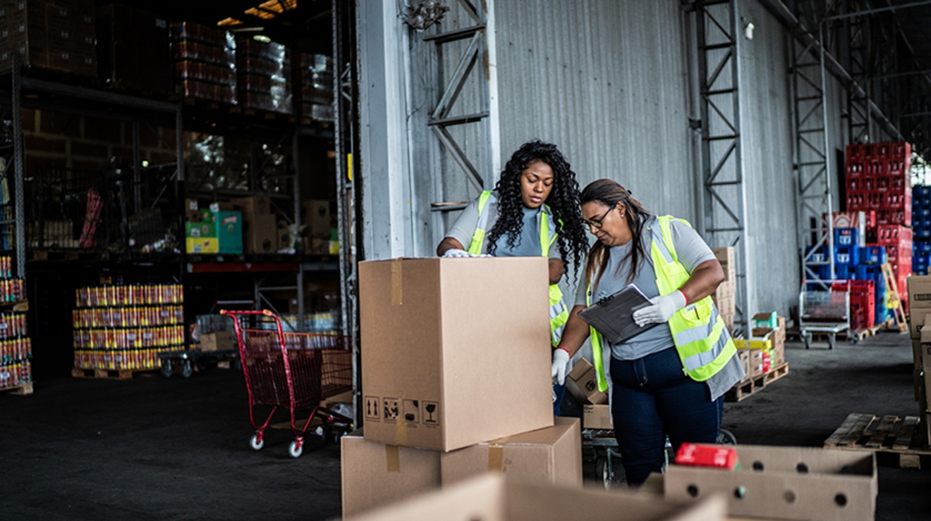 Two women wearing yellow vests stand in a factory looking at a large box