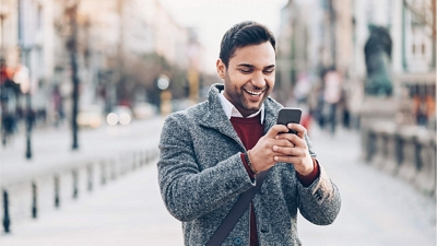 A smiling professional checks his smartphone while stopped on a city sidewalk.