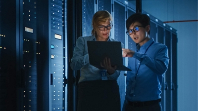 Two technicians review information on a laptop while standing in a server room.