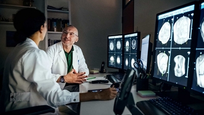 Two clinicians discuss medical data as they review images on multiple computer monitors.