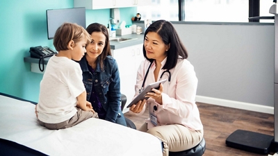 A pediatric doctor shows information on a tablet to a patient and his mother.