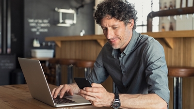 A salesperson uses T-Mobile for Microsoft Teams on a laptop and phone in a coffee shop.