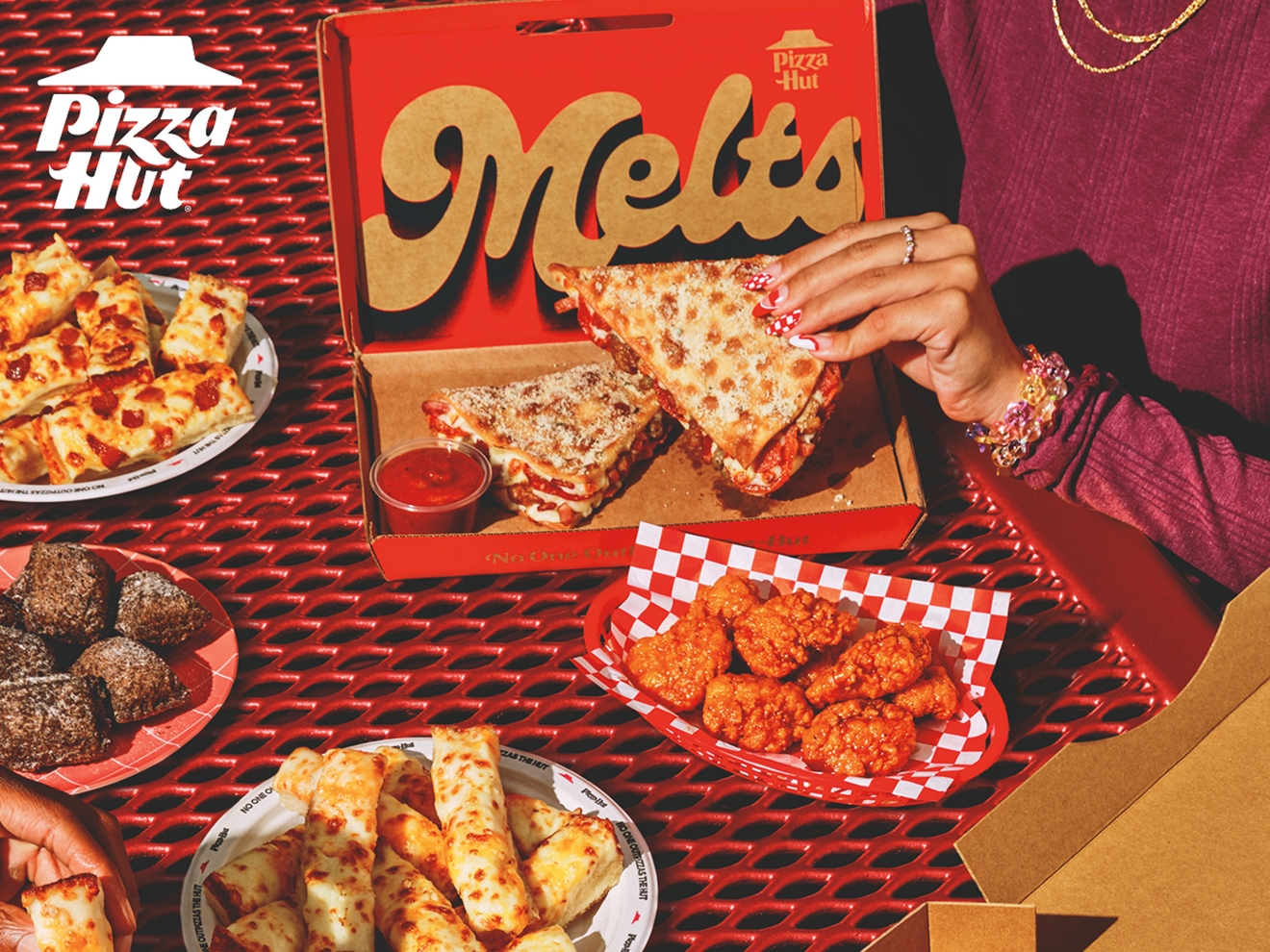 A hand holding a Pizza Hut Melt with wings, breadsticks, and desserts on a red picnic table.