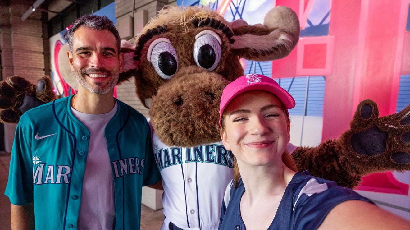 Two friends at a baseball game pose for a picture with the Mariners Moose.