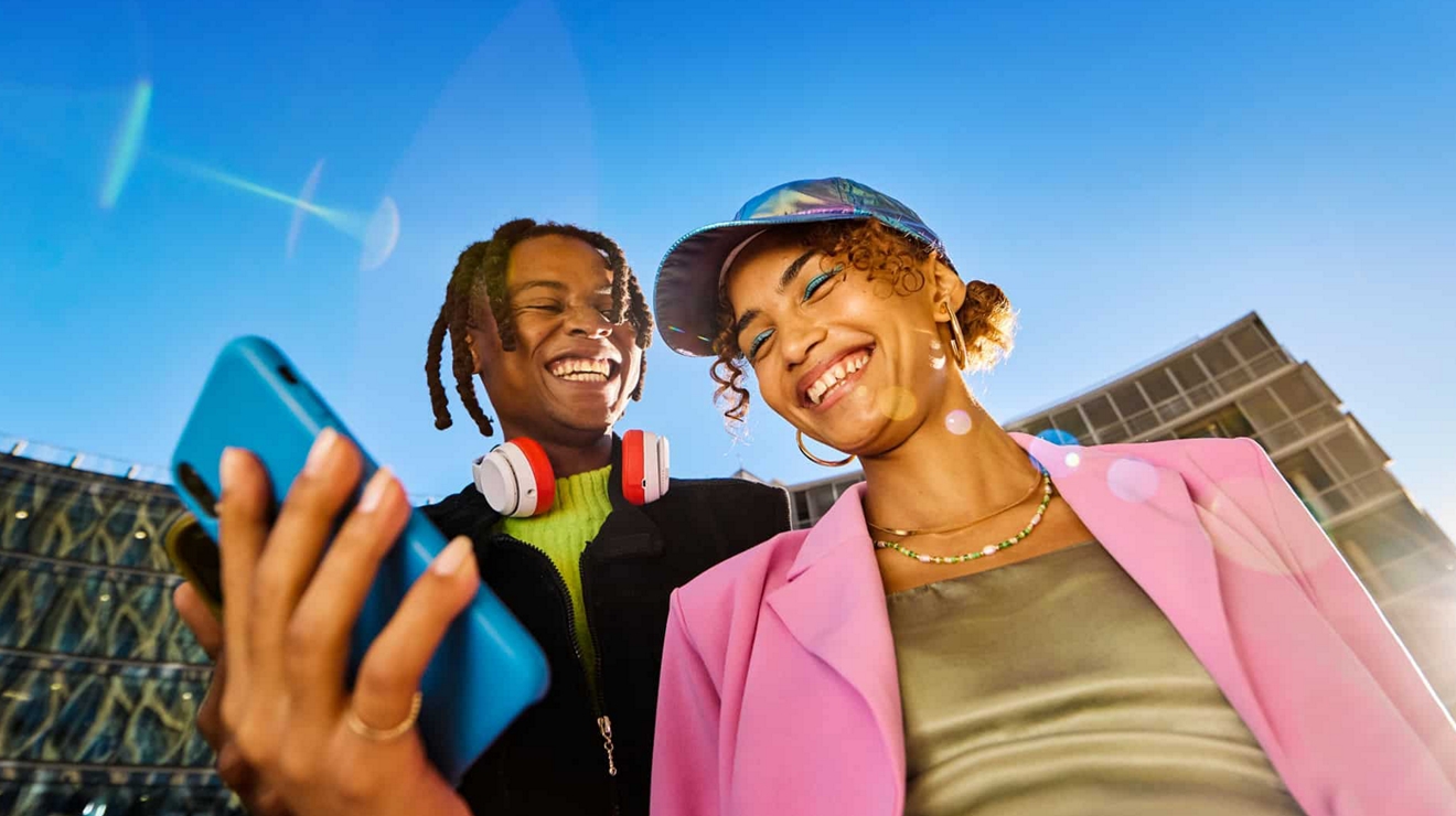 Couple laughing while looking at a device, with building and blue sky in the background.