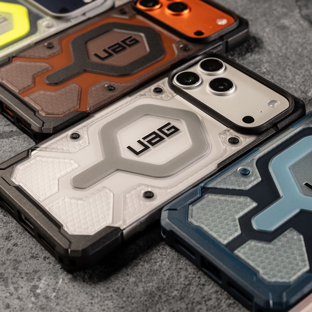 Four iPhone Pro Max’s in rugged UAG cases in colors silver, orange, and blue are arranged together, showcasing reinforced corners, textured designs, and raised camera protection