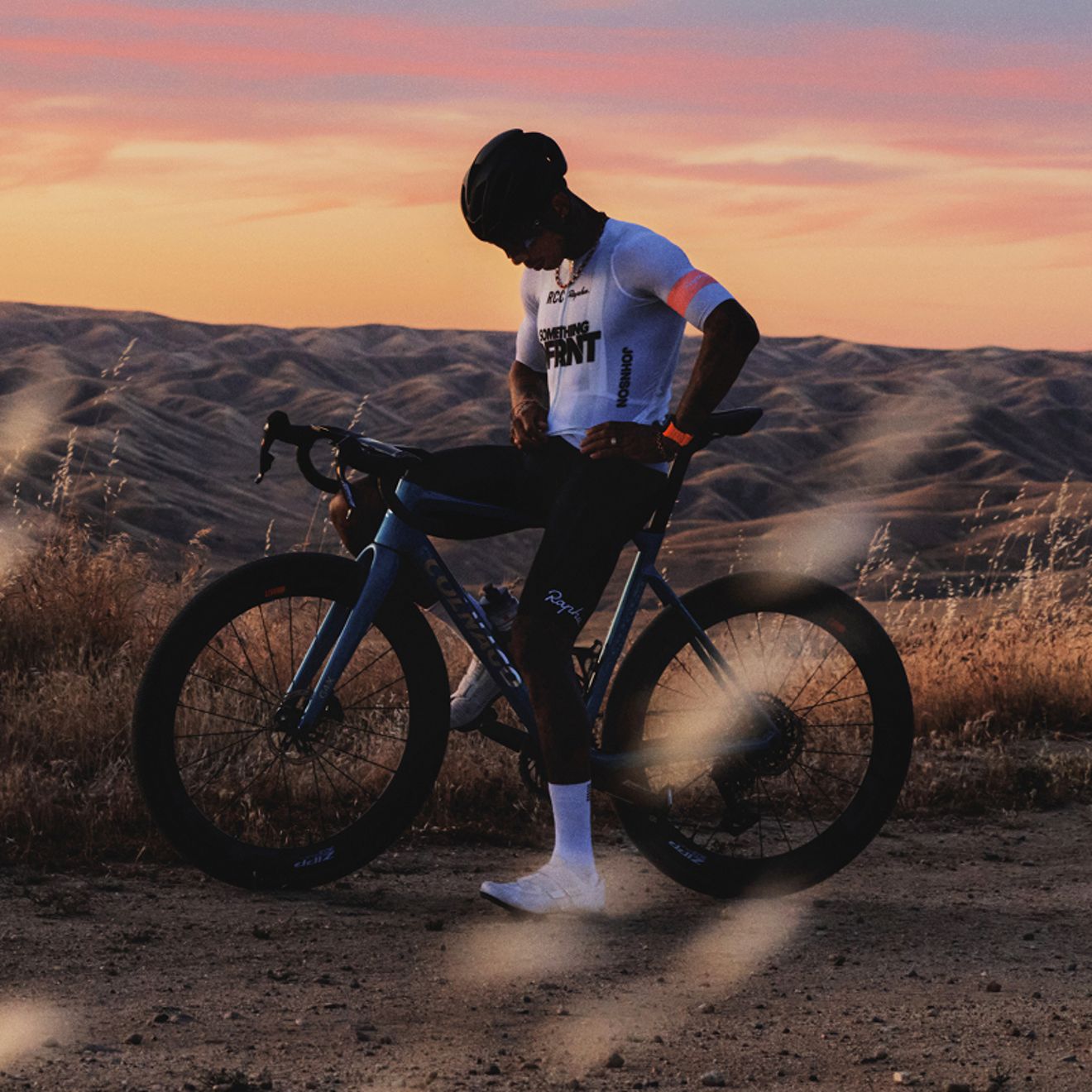 A cyclist wearing a helmet and cycling gear stands beside a road bike on a dirt trail, with rolling hills and a colorful sunset in the background.