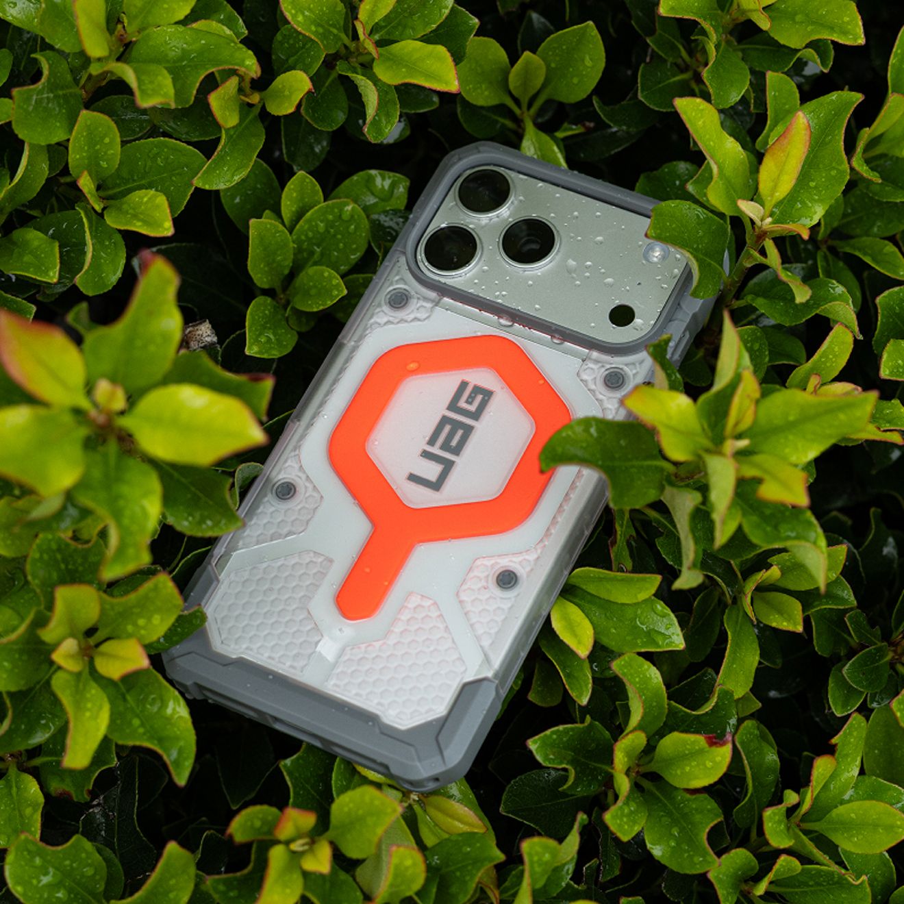 A smartphone in a rugged white and gray UAG case with bright orange accents rests among wet green leaves, with water droplets visible on the phone and case.