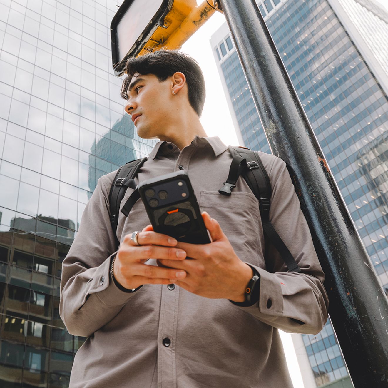 A person wearing a backpack holds a smartphone in a rugged UAG case while standing among tall buildings in a city.