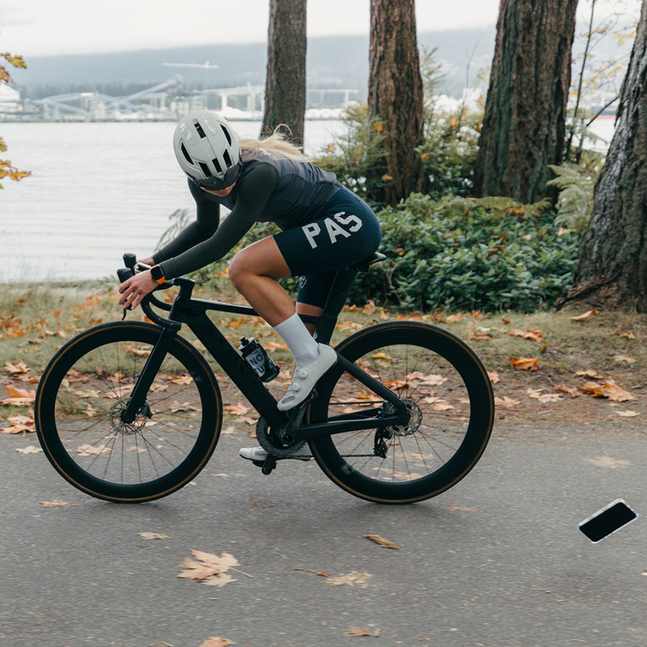 A cyclist wearing a helmet and black cycling gear rides a road bike along a waterfront path as a smartphone falls toward the pavement nearby.