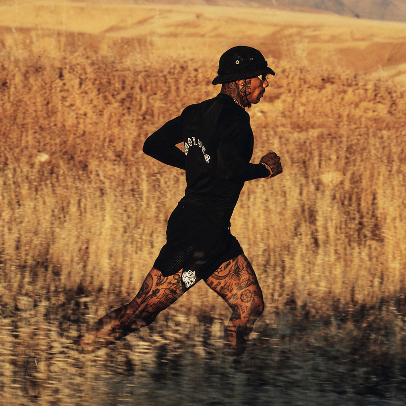 A tattooed runner wearing black athletic gear, a bucket hat, and sunglasses runs through a sunlit field.