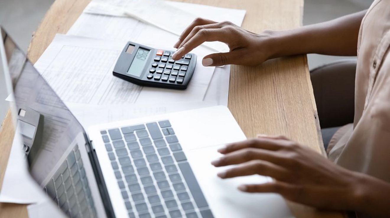 A person is sitting at a desk, typing on a laptop keyboard and calculator.